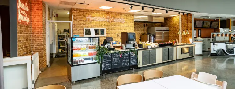 Kahvila-kiosk sisätiloissa tiiliseinien ympäröimänä, myyntitiski kahvikoneineen sekä lasiovissa jäätelö- ja mehuvalikoima.