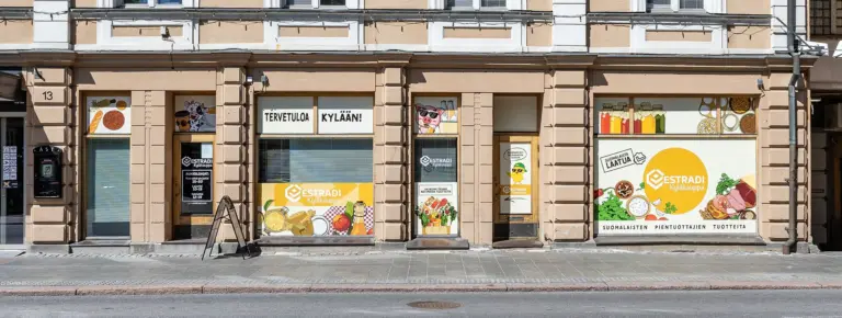 Estradi Kyläkauppa elintarvikeliike vanhassa kivitalossa; näyteikkuna täynnä ruokatarvikkeita ja Tervetuloa kylään -viesti.