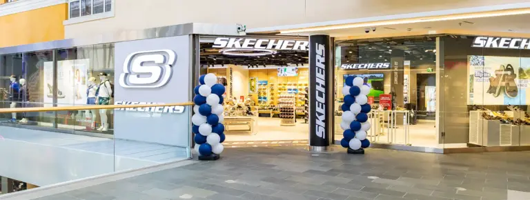 Skechers-kauppa ostoskeskuksessa, kirkkaasti valaistu sisäänkäynti ja sinivalkoiset ilmapallot ulko-ovien edessä.
