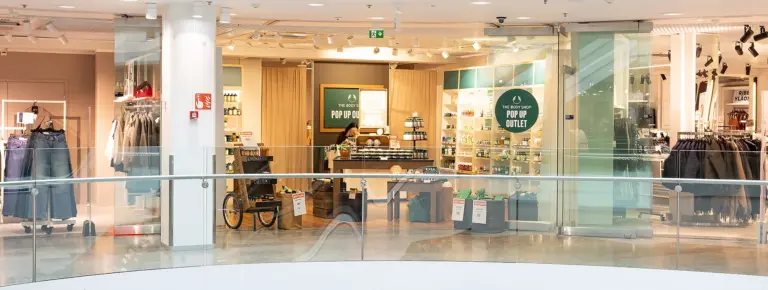 The Body Shop Pop Up Outlet -myymälä ostoskeskuksessa, vaatteita ja kosmetiikkaa esillä lasikaiteiden ympäröimässä tilassa.