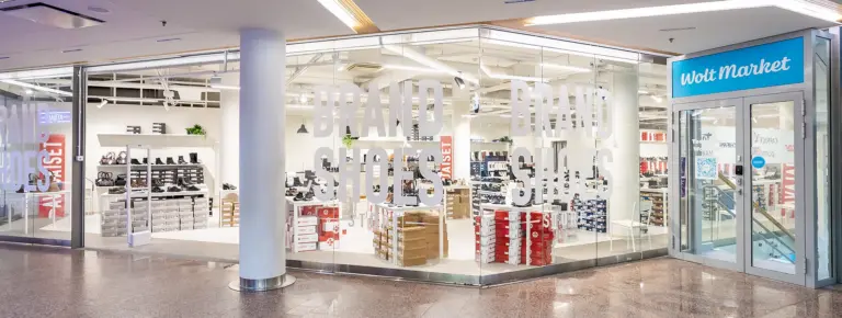 Lasiseinäinen kenkämyymälä BRAND SHOES -tekstillä, sisällä kiertävät hyllyt täynnä kenkiä; oikealla Wolt Marketin ovi.