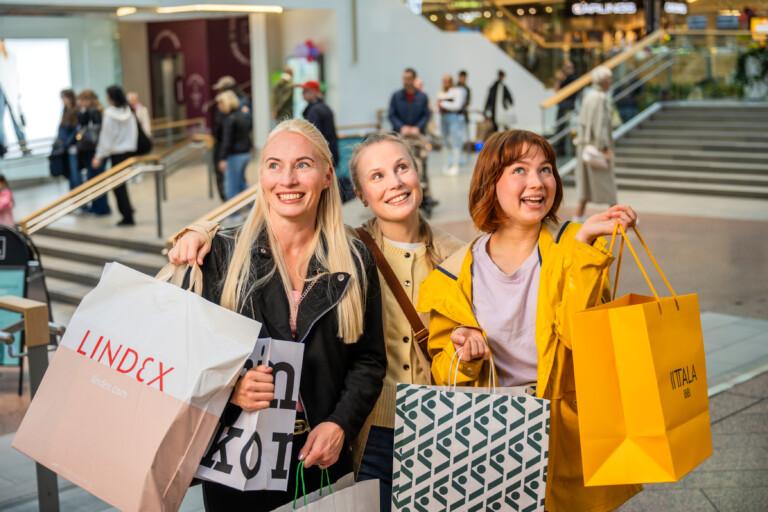 Kolme iloisesti hymyilevää naista ostoksilla kauppakeskuksessa, kantavat värikkäitä ostoskasseja ja naurattavat toisiaan.